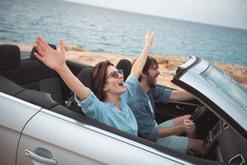 Couple rides in beach in convertible with top down, woman's hands thrown up wide